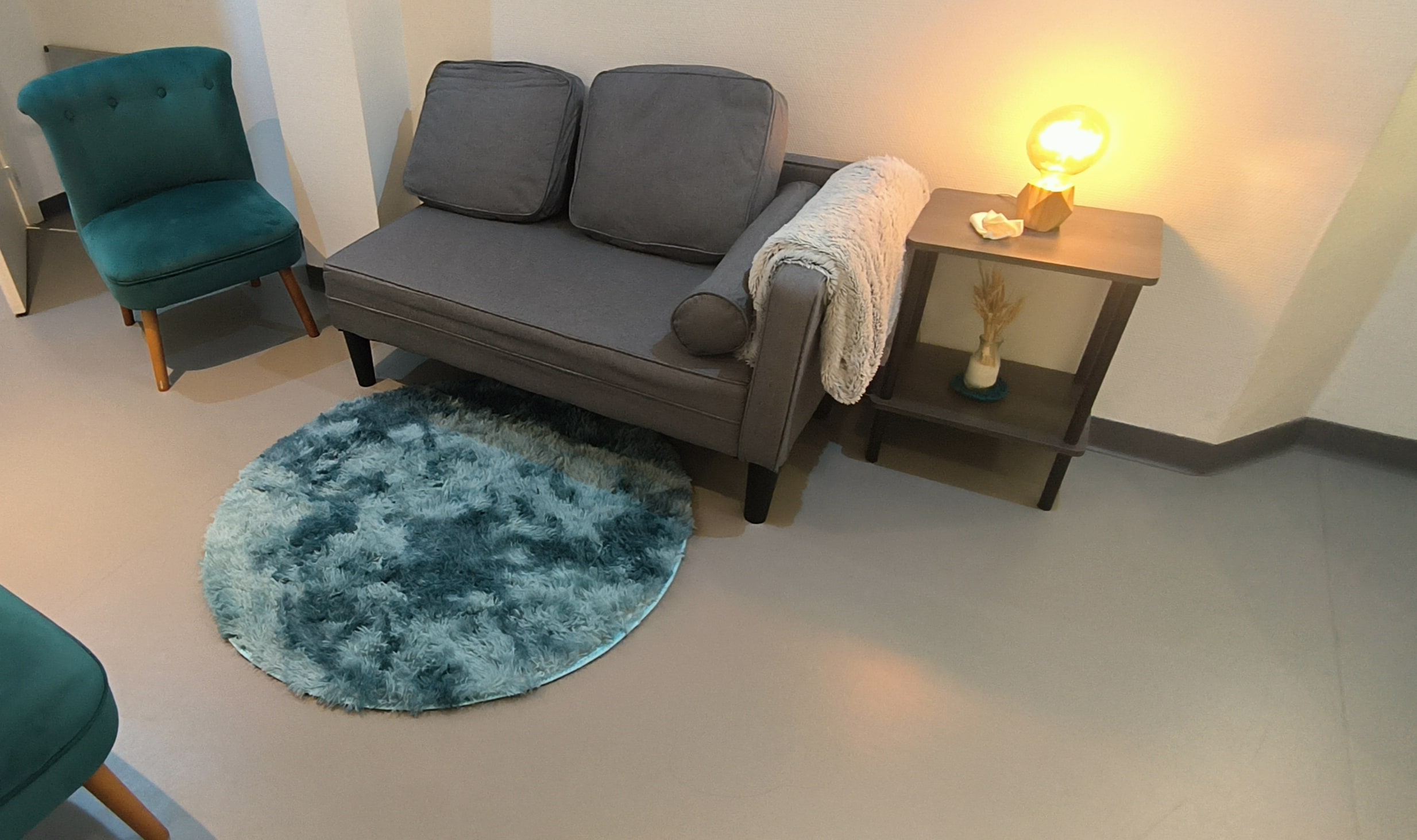 Espace de consultation du cabinet de psychologue à Mandelieu-La Napoule, avec canapé gris, fauteuil vert et tapis rond sous une lampe d’ambiance.